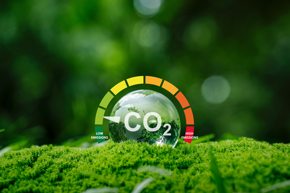 CO₂-Emissionsskala mit Zeiger im grünen Bereich, davor eine Glaskugel mit Waldspiegelung auf Moos