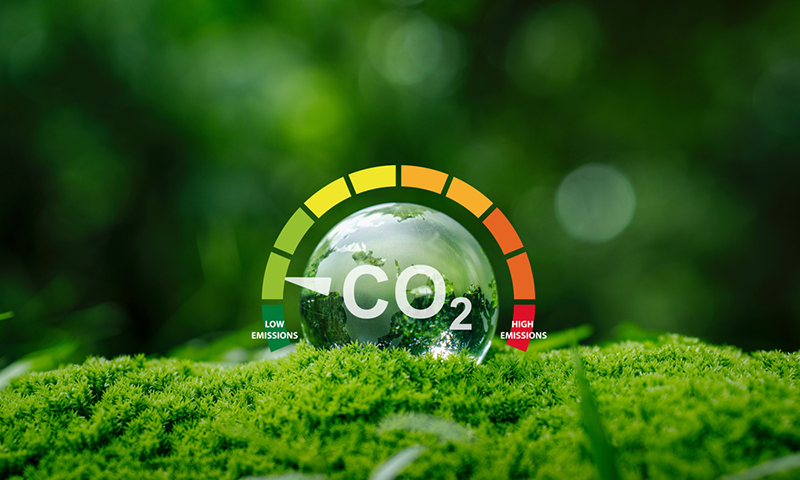 CO₂-Emissionsskala mit Zeiger im grünen Bereich, davor eine Glaskugel mit Waldspiegelung auf Moos